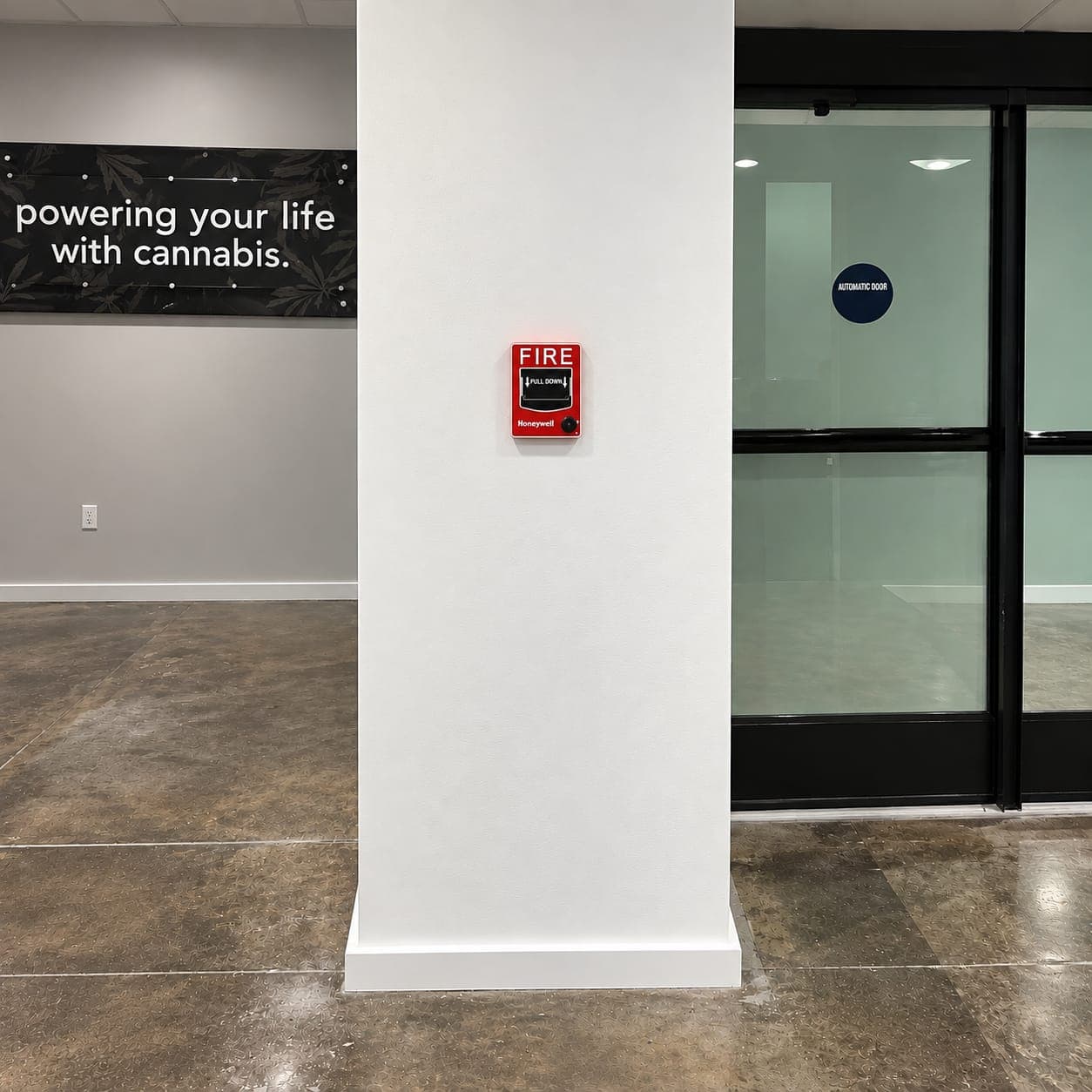Square column wrapped in smooth white drywall with base trim and a fire alarm pull station, beside glass entry doors