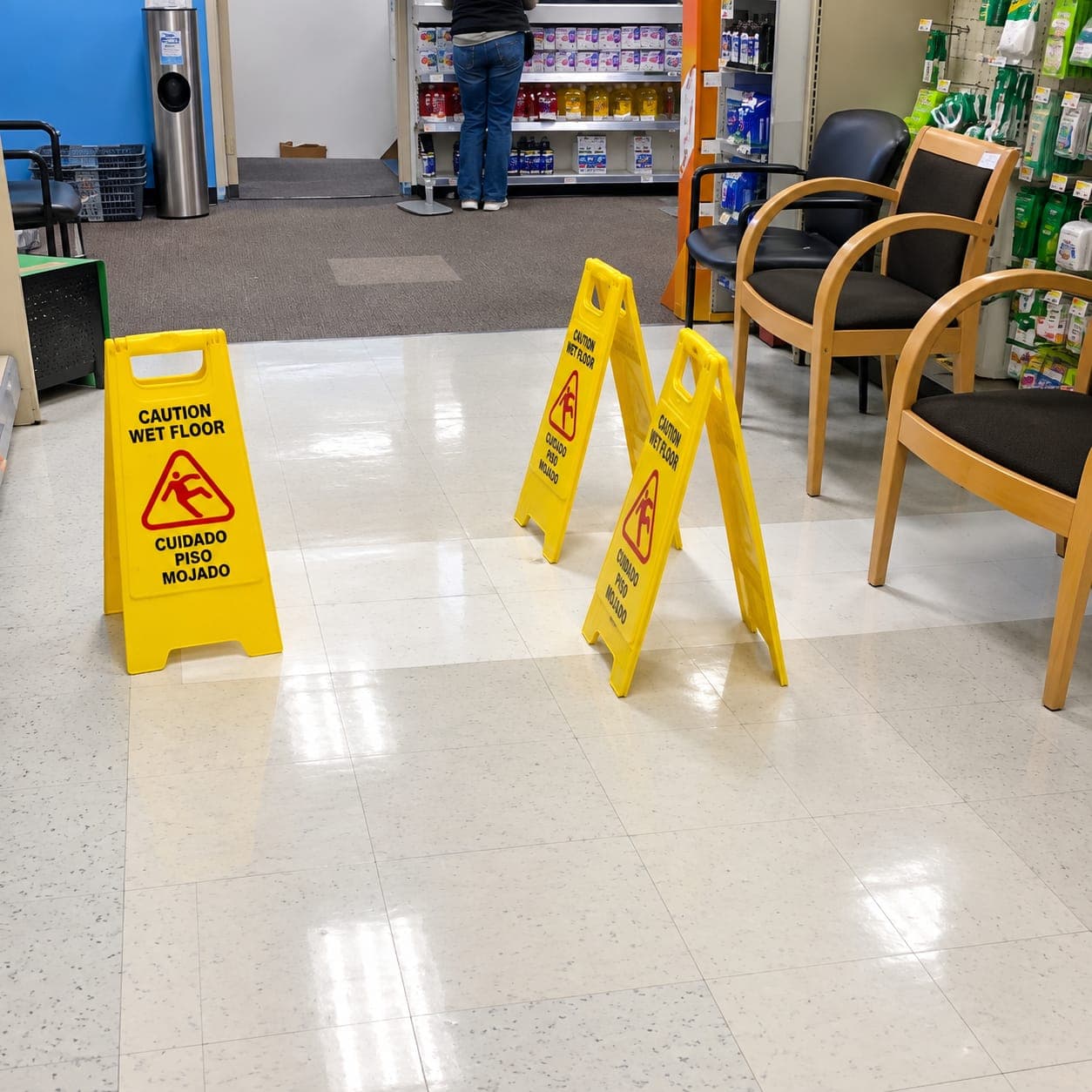 Glossy white speckled vinyl floor tiles in a store waiting area with three yellow wet floor signs and wooden chairs