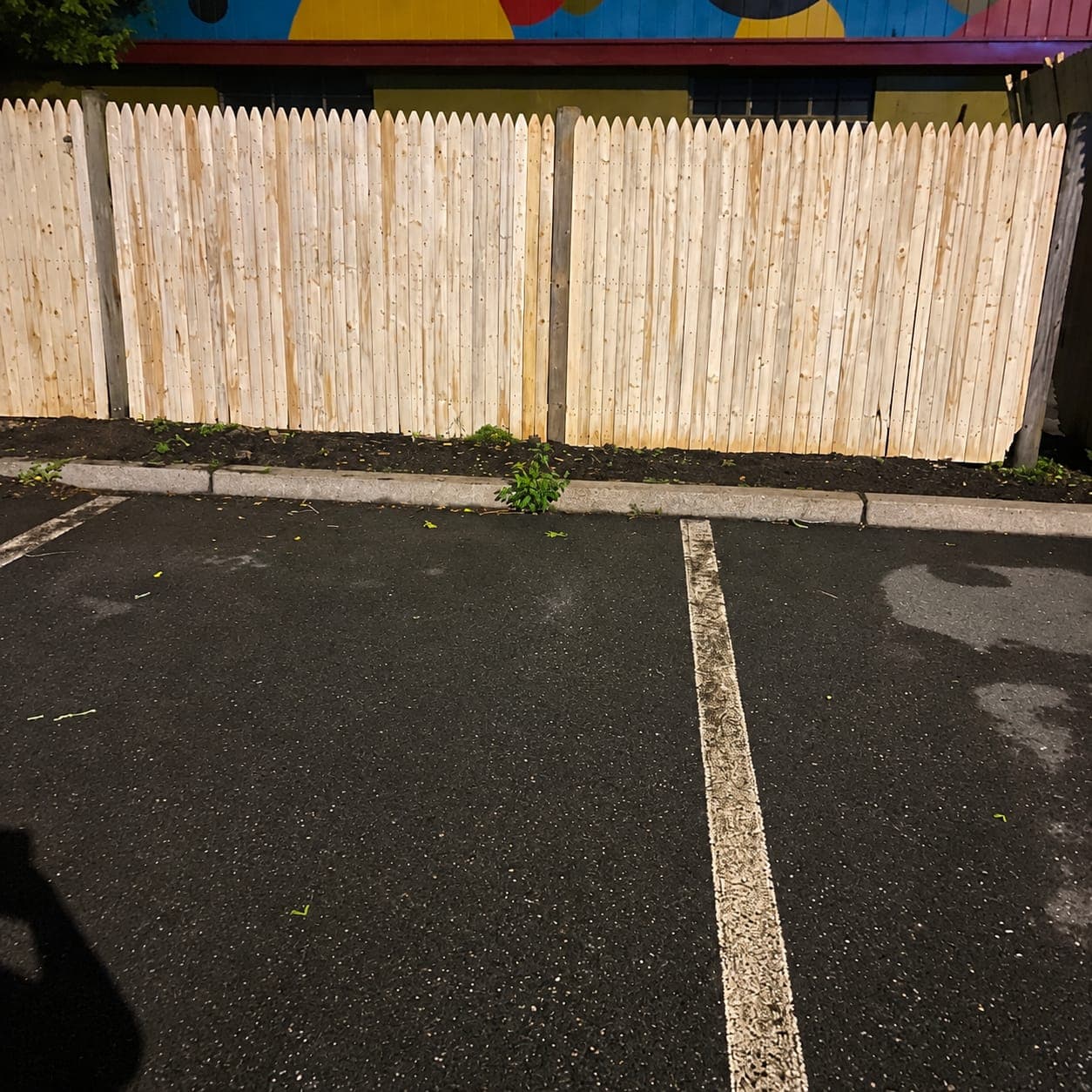 New pointed-picket stockade fence panels on weathered posts behind a parking lot curb, with a painted mural above