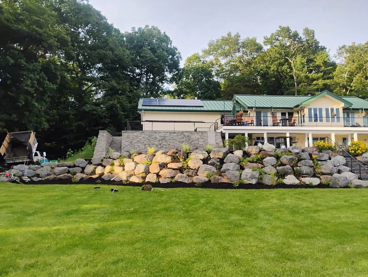 Boulder retaining wall with new plantings and fresh black mulch below a green-roofed home and stone steps