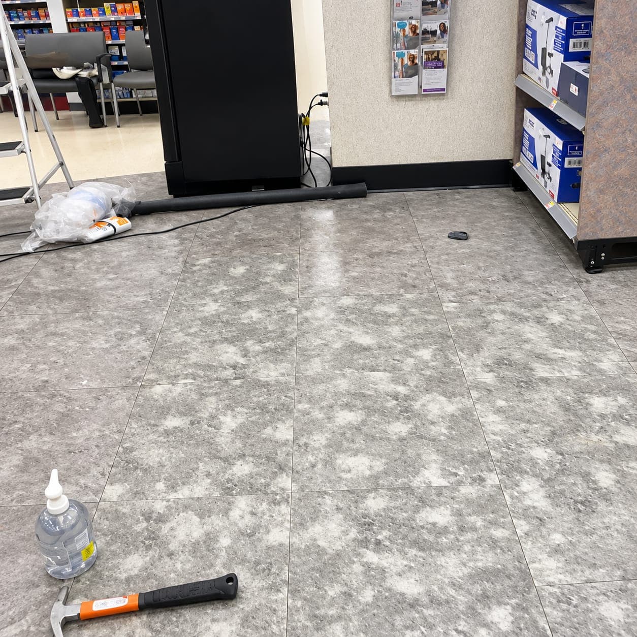 Newly laid gray mottled floor tiles in a retail store, with a hammer, squeeze bottle and step ladder nearby