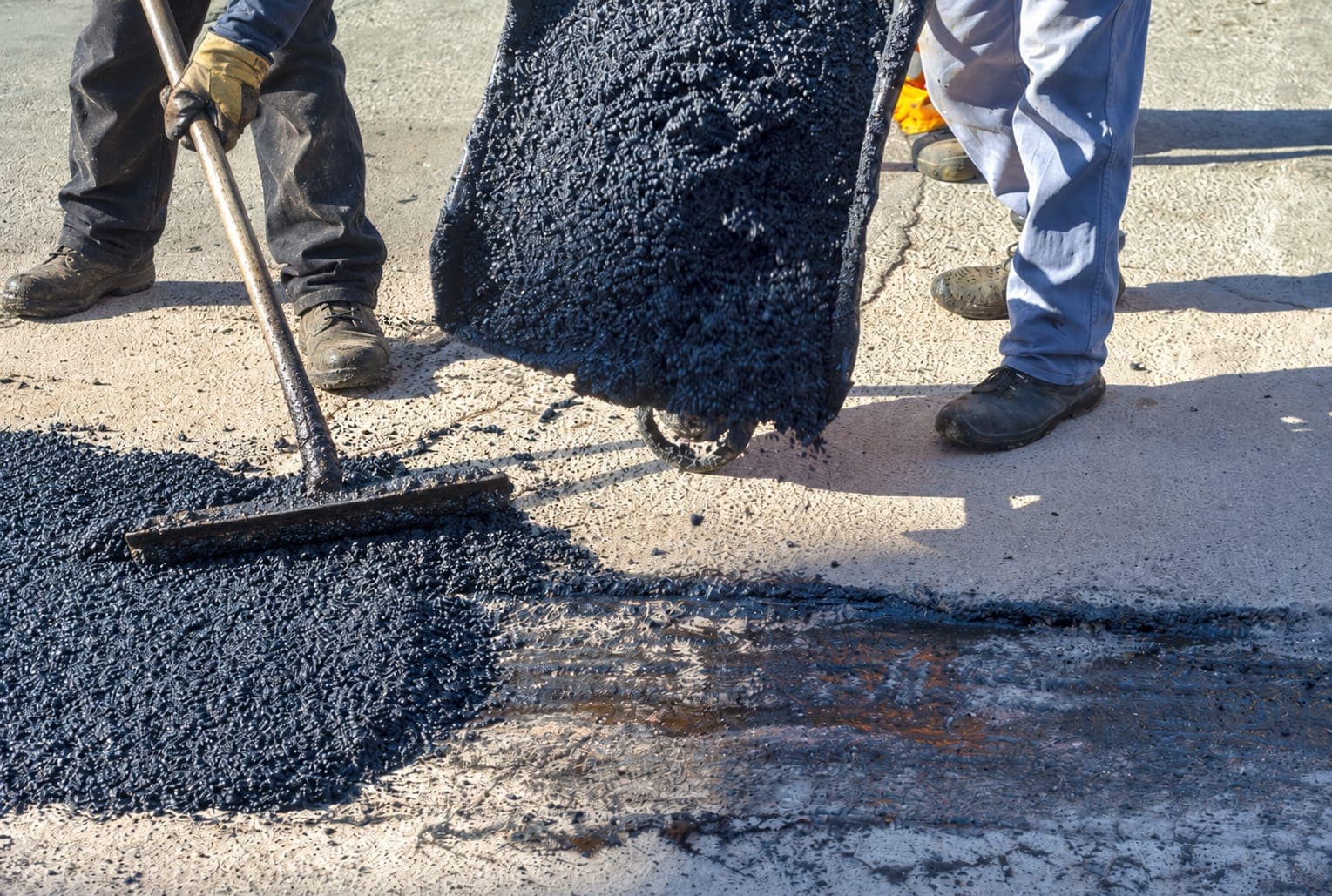 Workers dumping asphalt from a wheelbarrow and spreading it with a rake to patch damaged pavement