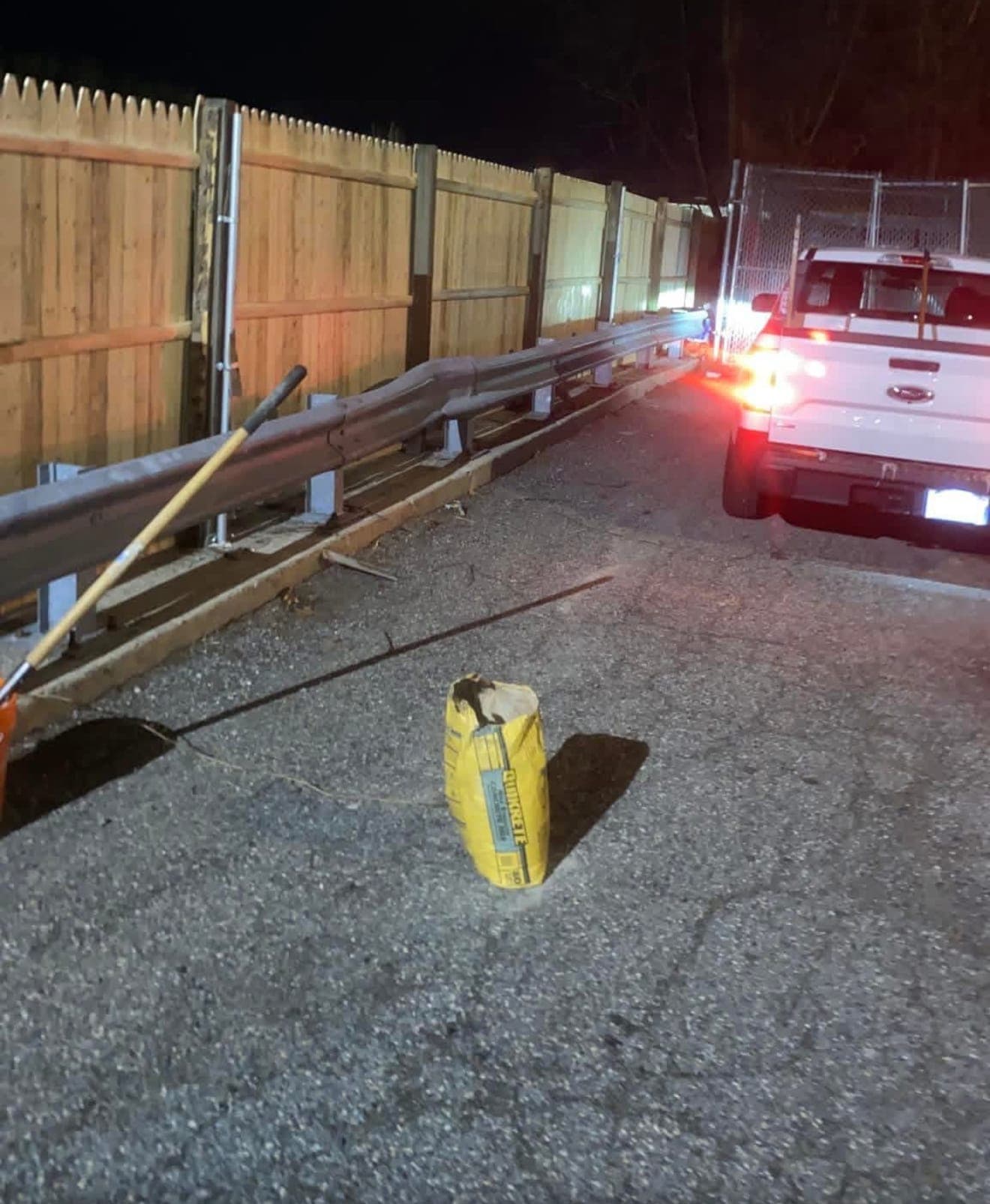 Wood stockade fence and steel guardrail along a parking lot at night, with a shovel and bag of concrete mix