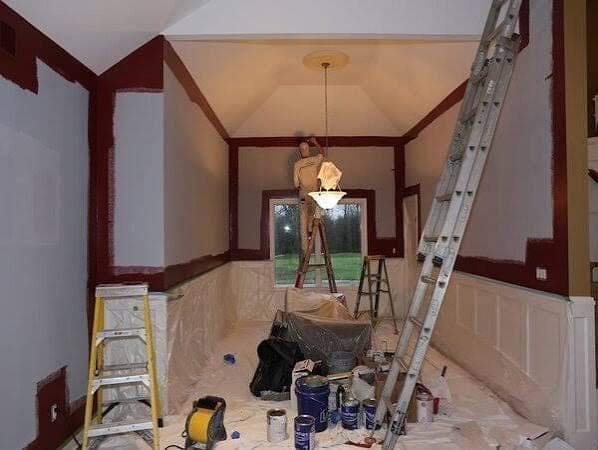 Interior repaint in progress with walls part red and part gray, drop cloths, ladders and a painter near the ceiling