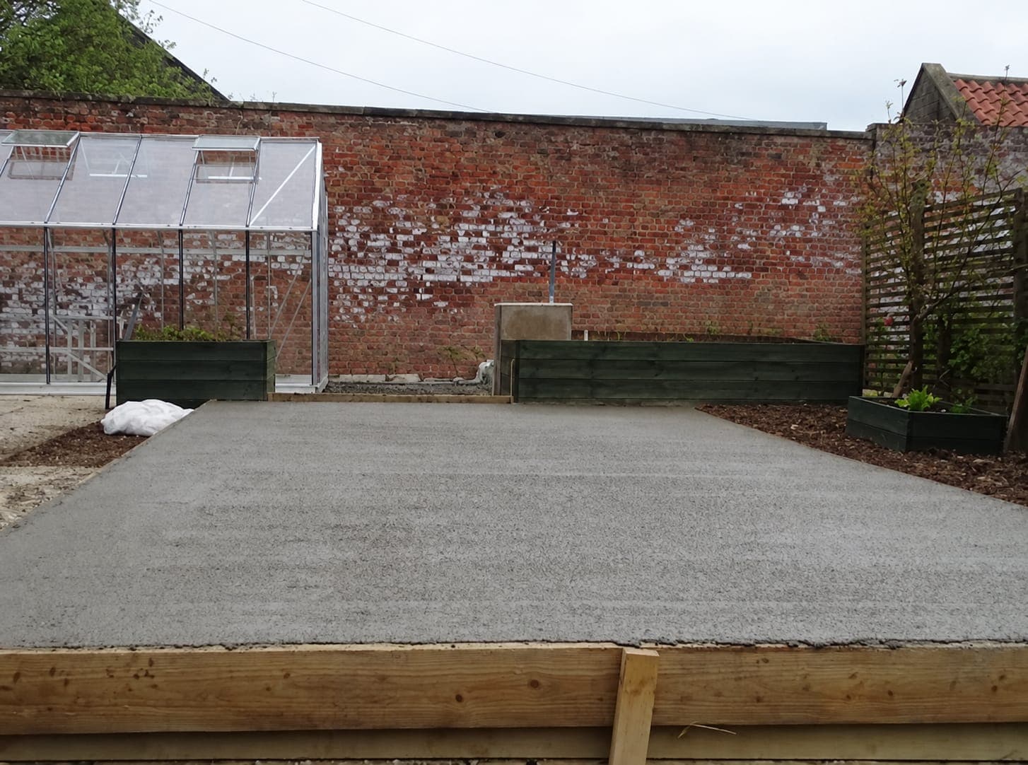 New concrete pad with a ridged finish inside timber forms, in a walled garden beside a greenhouse and raised beds