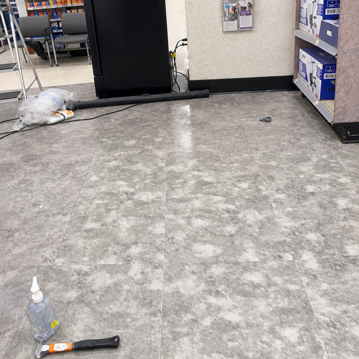Gray mottled stone-look floor tiles in a retail store, with a floor scraper, squeeze bottle and step ladder on site