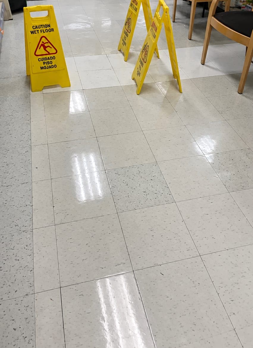 Glossy cream speckled floor tiles in a commercial space, with yellow wet floor signs and wooden chairs nearby