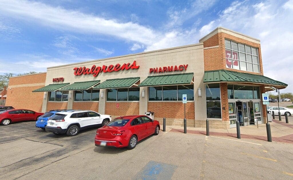 Brick retail pharmacy with green metal awnings and red signage, cars parked in the asphalt lot out front