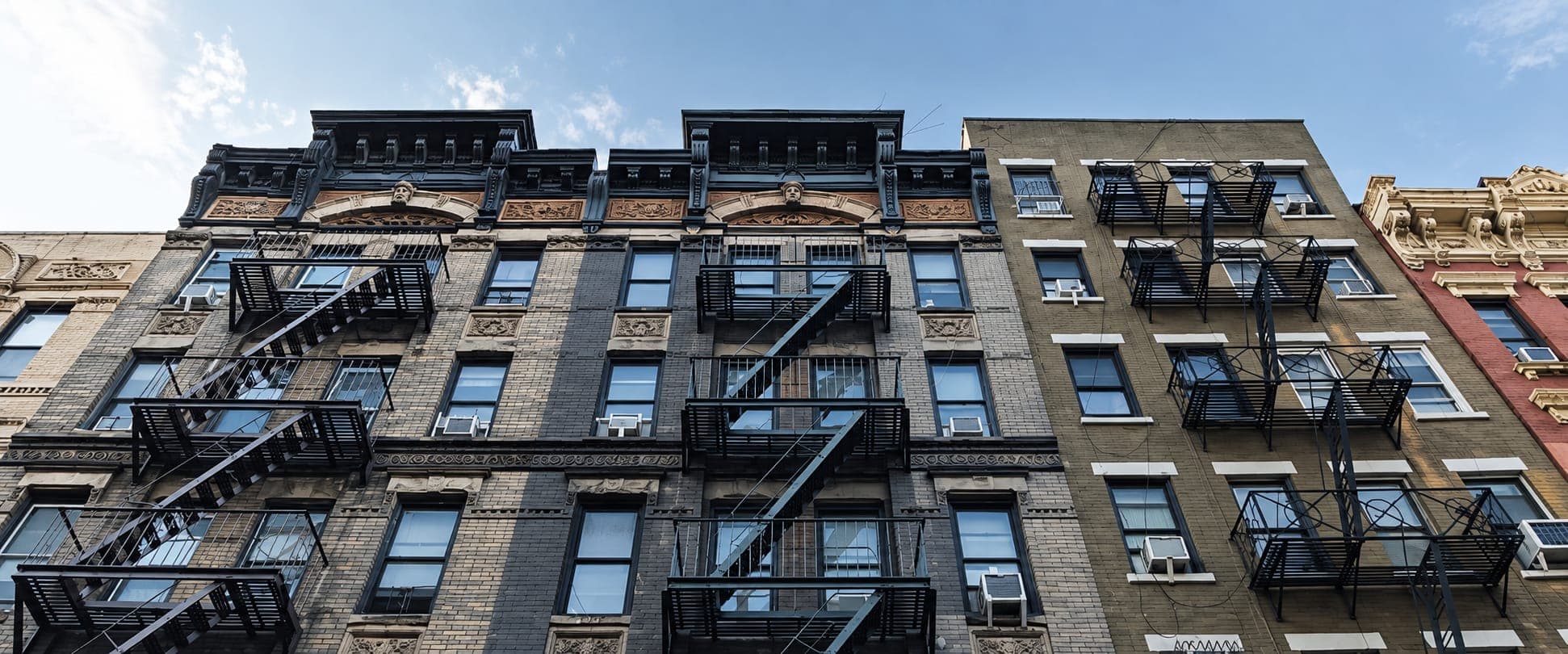 Looking up at older brick apartment buildings with black steel fire escapes, window air conditioners and ornate cornices