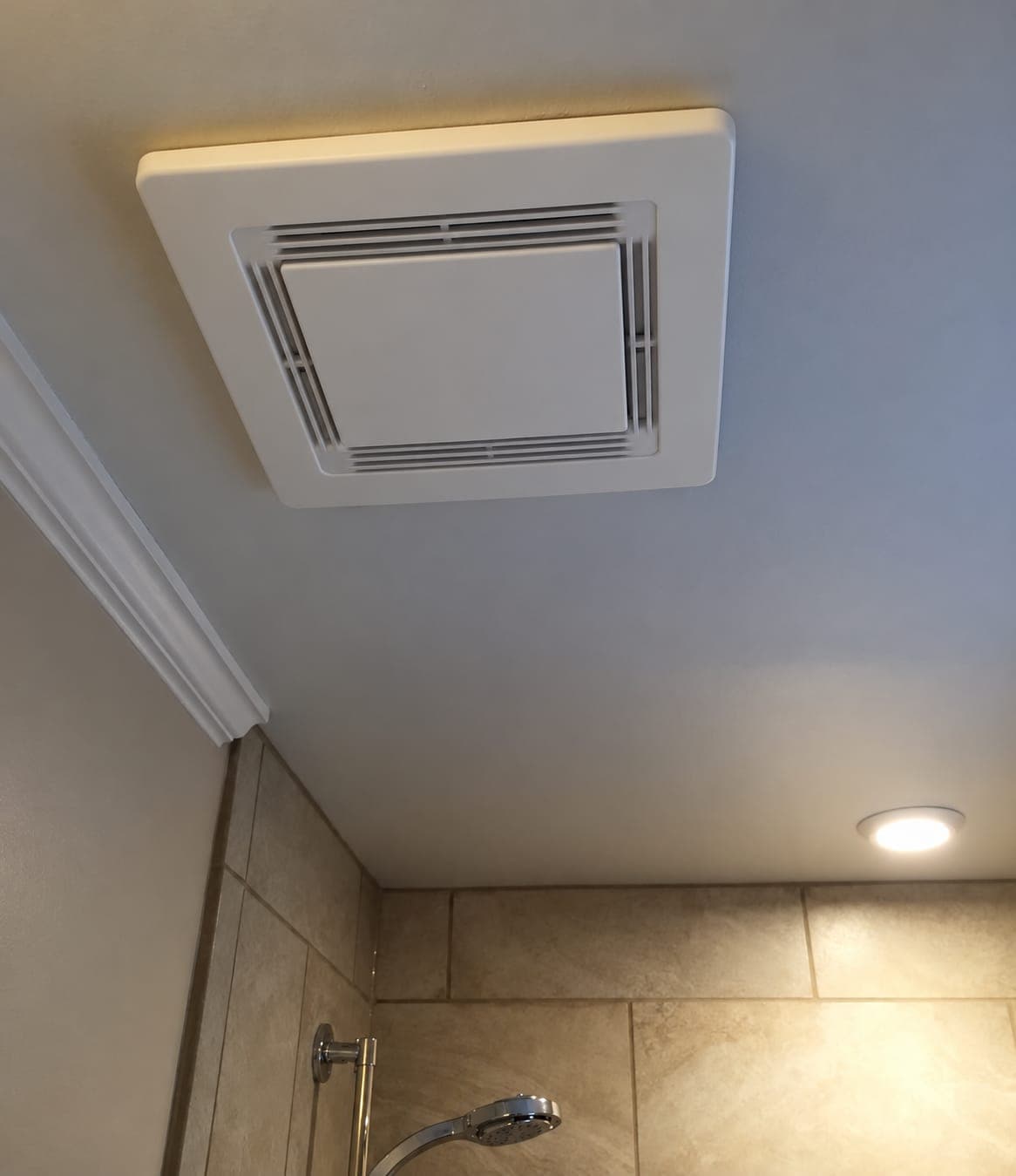 White square exhaust fan grille in a bathroom ceiling near a beige tiled shower with a chrome handheld showerhead