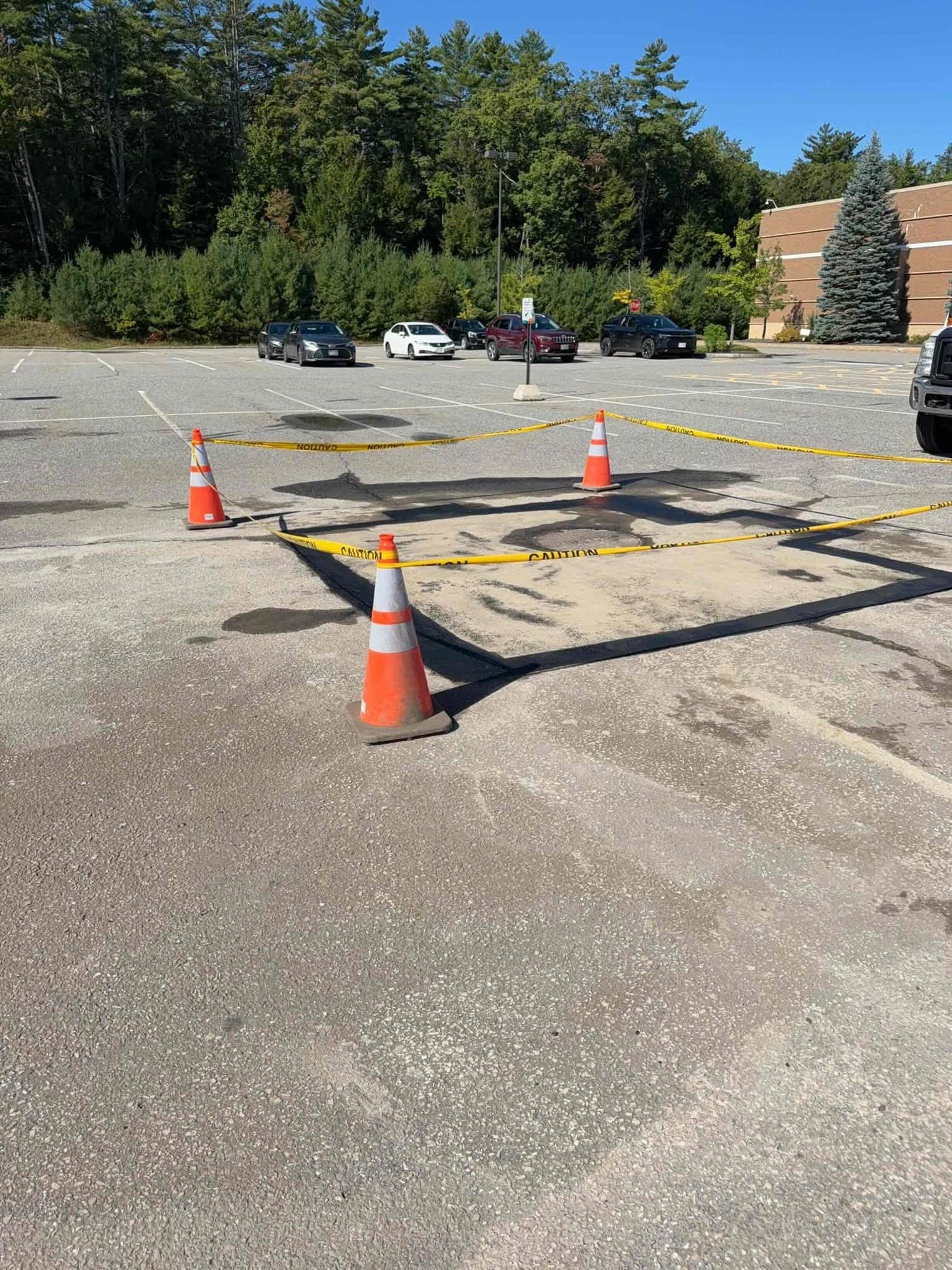 Square asphalt patch with sealed edges around a parking lot storm drain, roped off with cones and caution tape