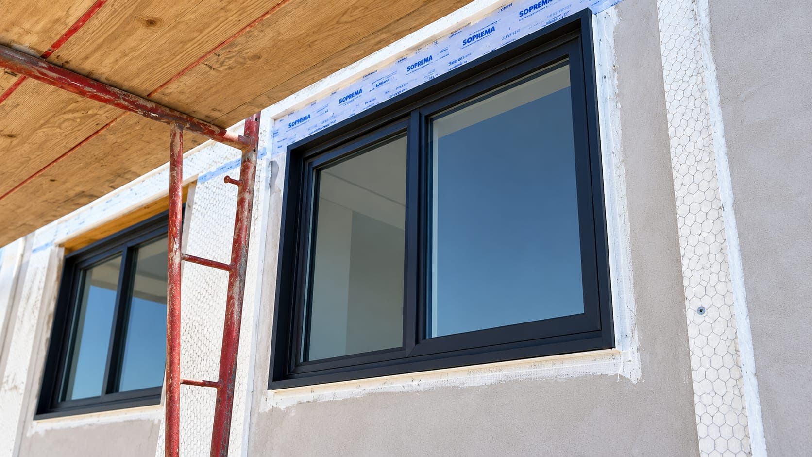 Newly installed black-framed sliding windows in a stucco wall under construction, beside red scaffolding