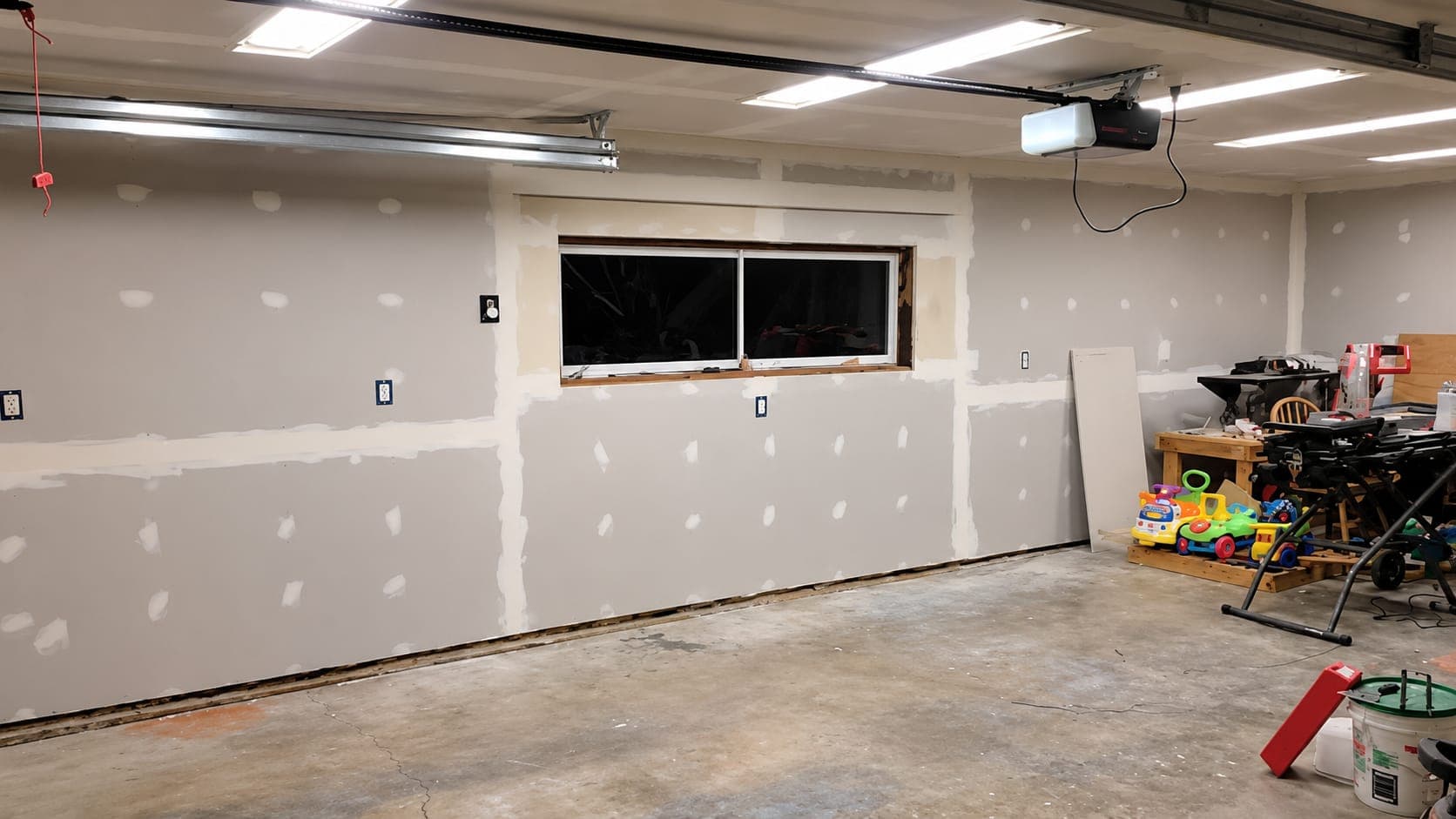 New gray drywall hung on garage walls, with taped seams and screw heads spotted in joint compound around a window