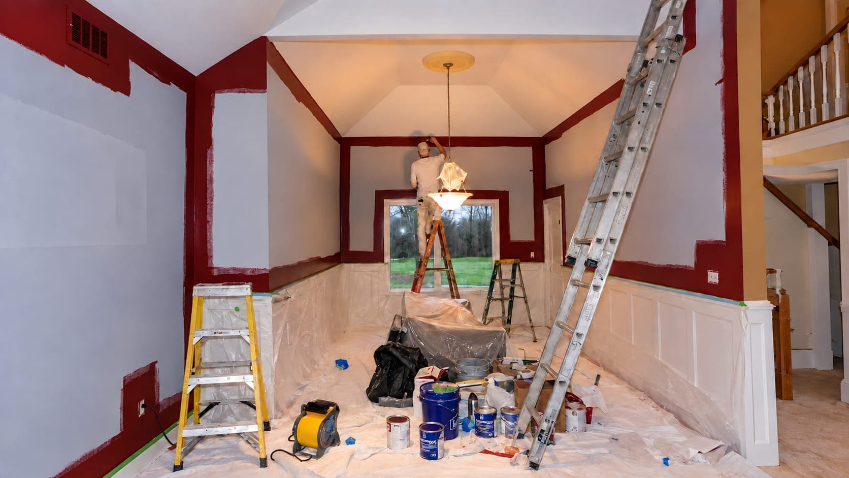 Painter on a stepladder cutting in along the ceiling of a deep red room being repainted pale gray