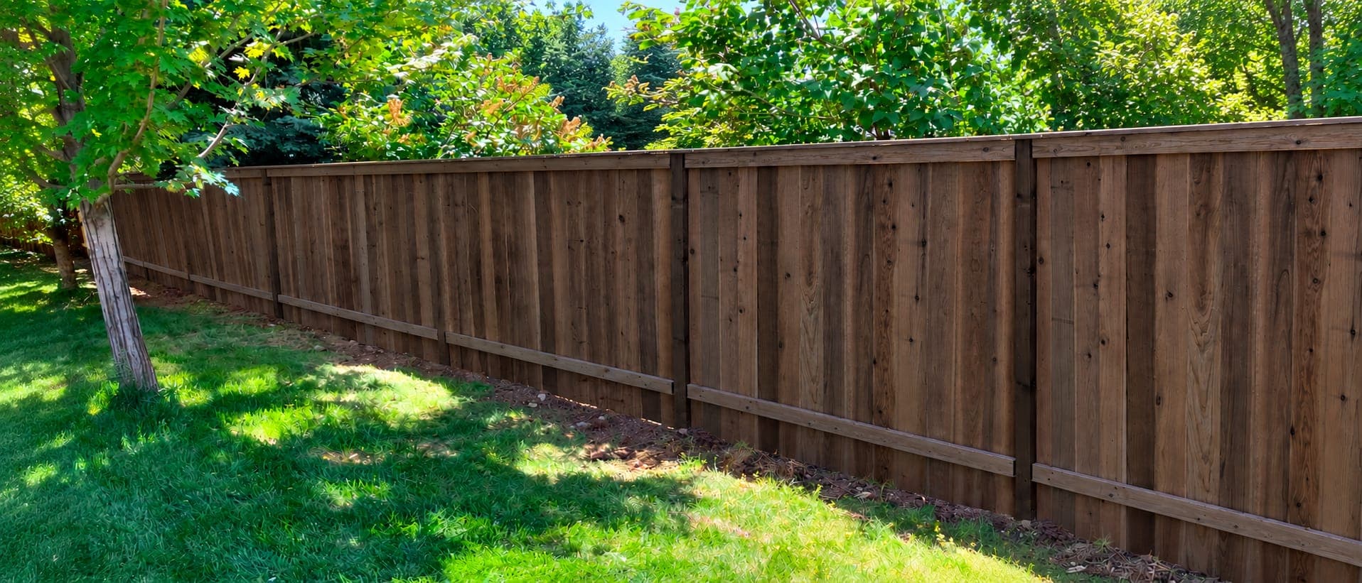 Long brown wood privacy fence with a flat top cap running along a mulched edge beside a sunny, tree-shaded lawn