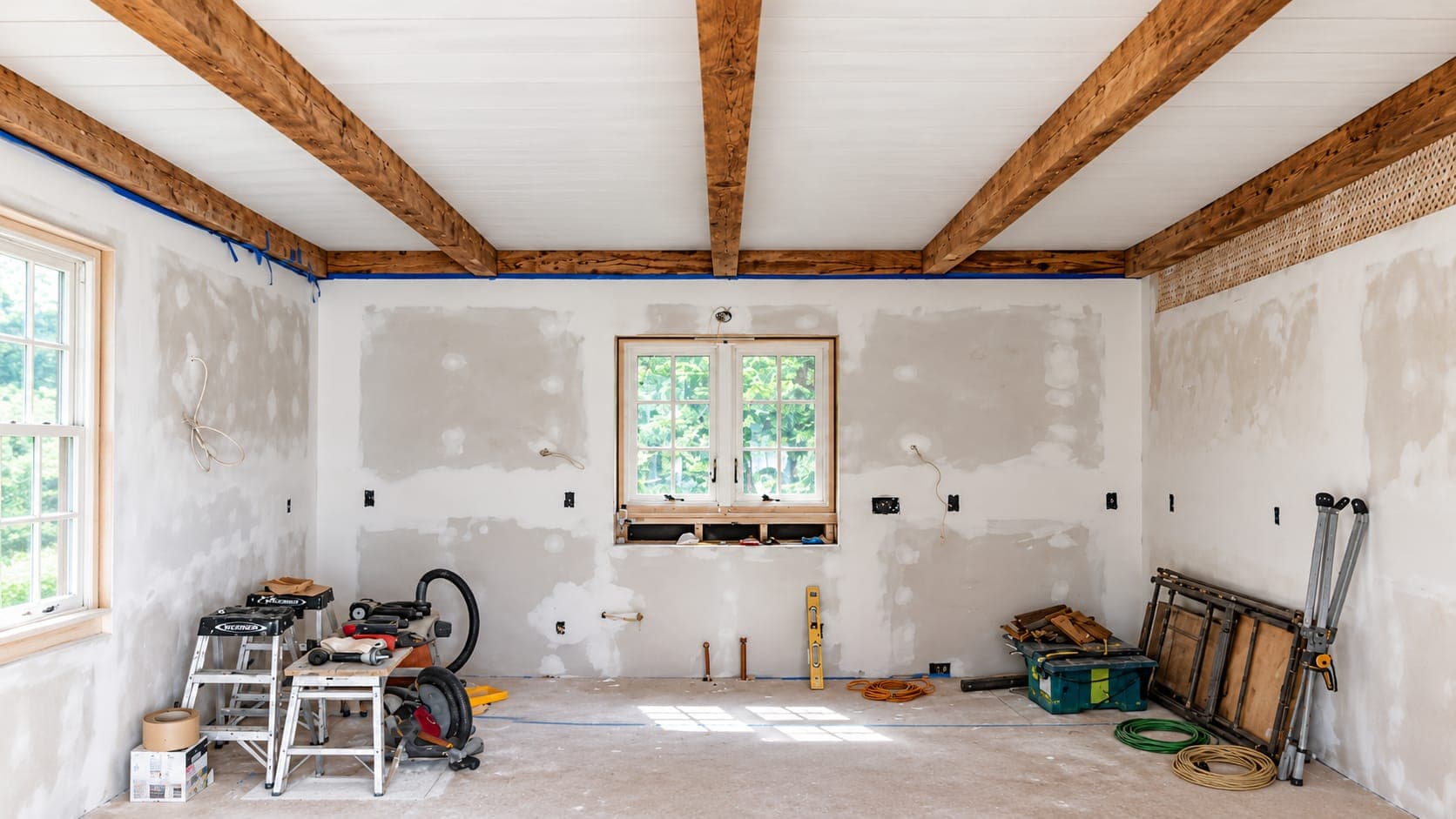 Room under renovation with patched drywall, rustic wood ceiling beams, an untrimmed window and tools on the floor