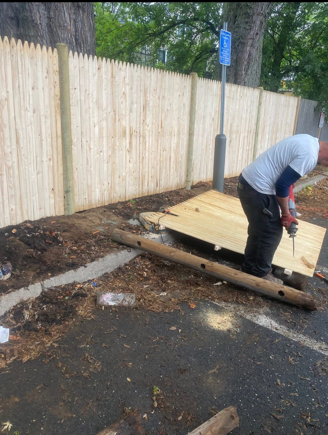 Worker cutting a wood stockade fence panel with a reciprocating saw beside a newly built fence in a parking lot