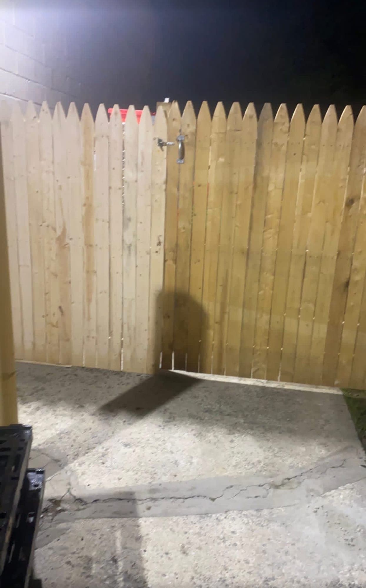 New wood stockade fence and gate with a latch and pull handle, set along a concrete pad and lit by floodlight at night