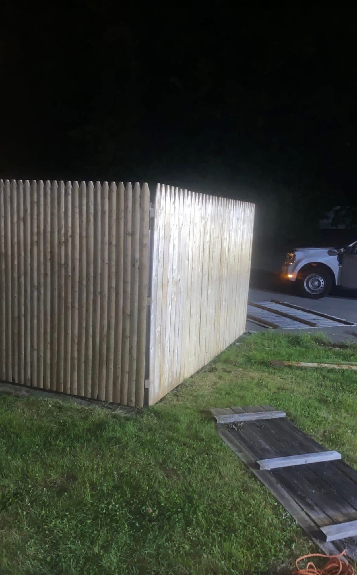Corner of a new wood stockade fence enclosure on a lawn at night, with old weathered fence panels lying on the grass