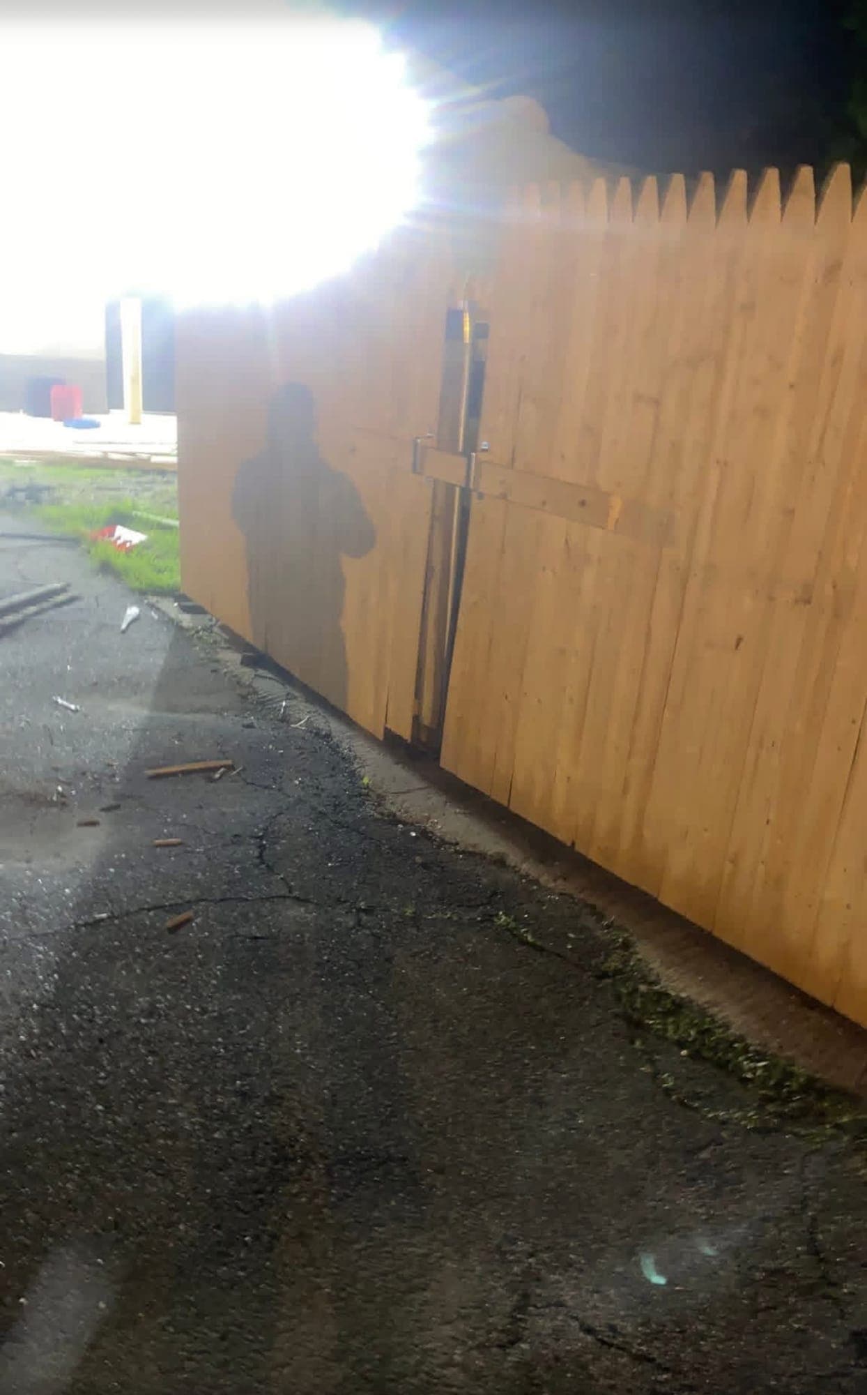 Wood stockade fence gate held shut by a wooden crossbar in metal brackets, beside cracked asphalt at night