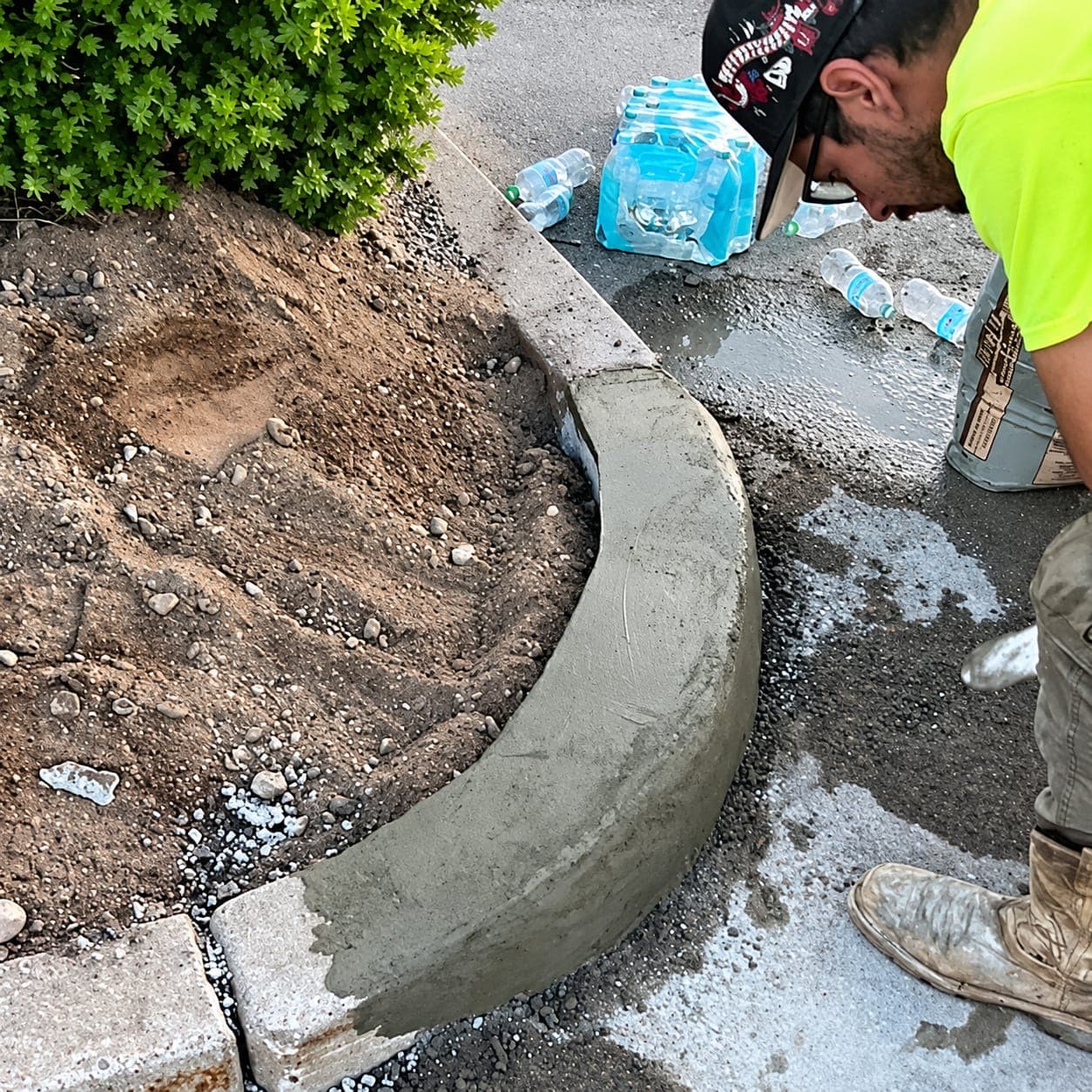 Worker in a hi-vis shirt finishing a fresh curved concrete curb along a landscape bed next to an asphalt parking lot