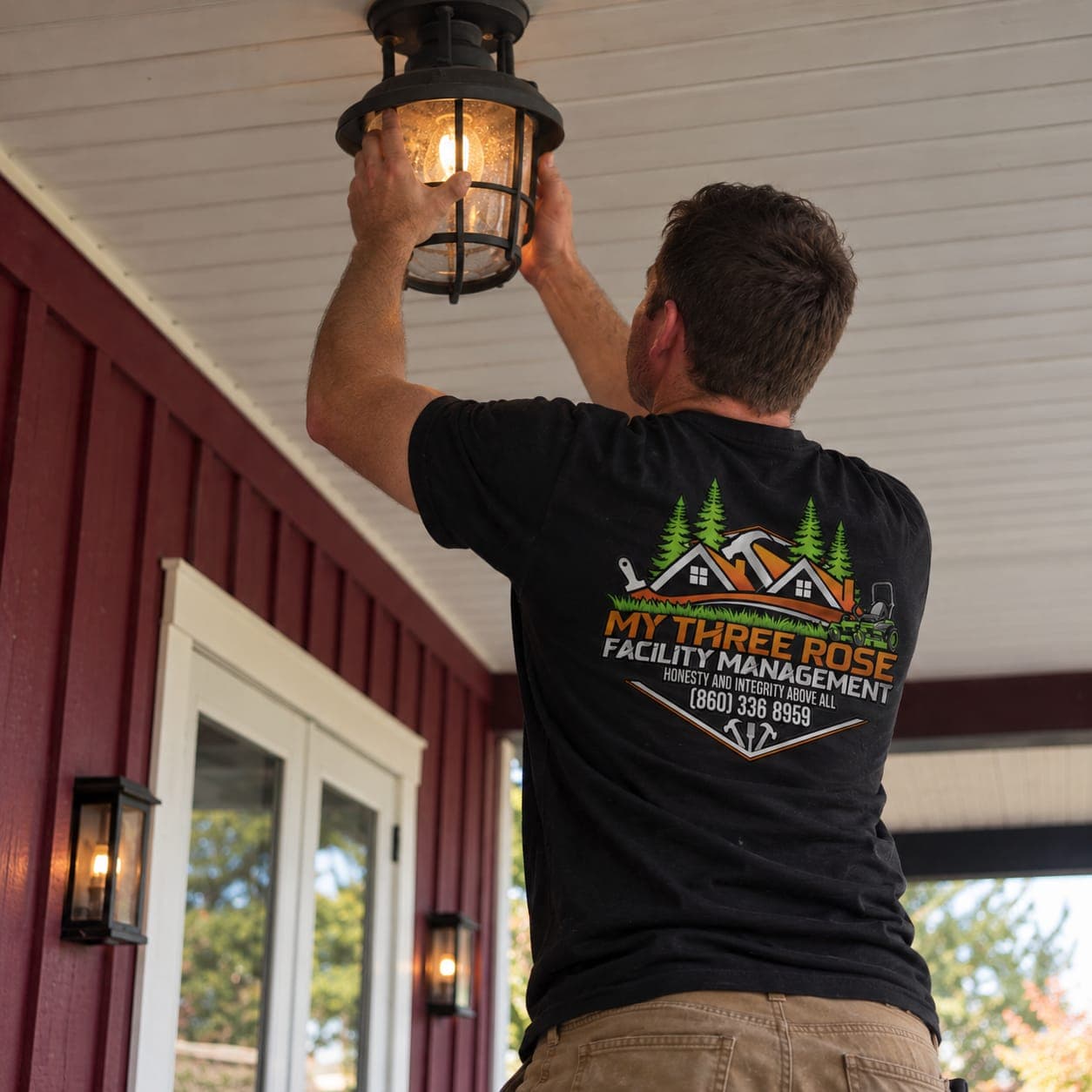 Handyman installing a black caged lantern light on a porch ceiling beside red board-and-batten siding
