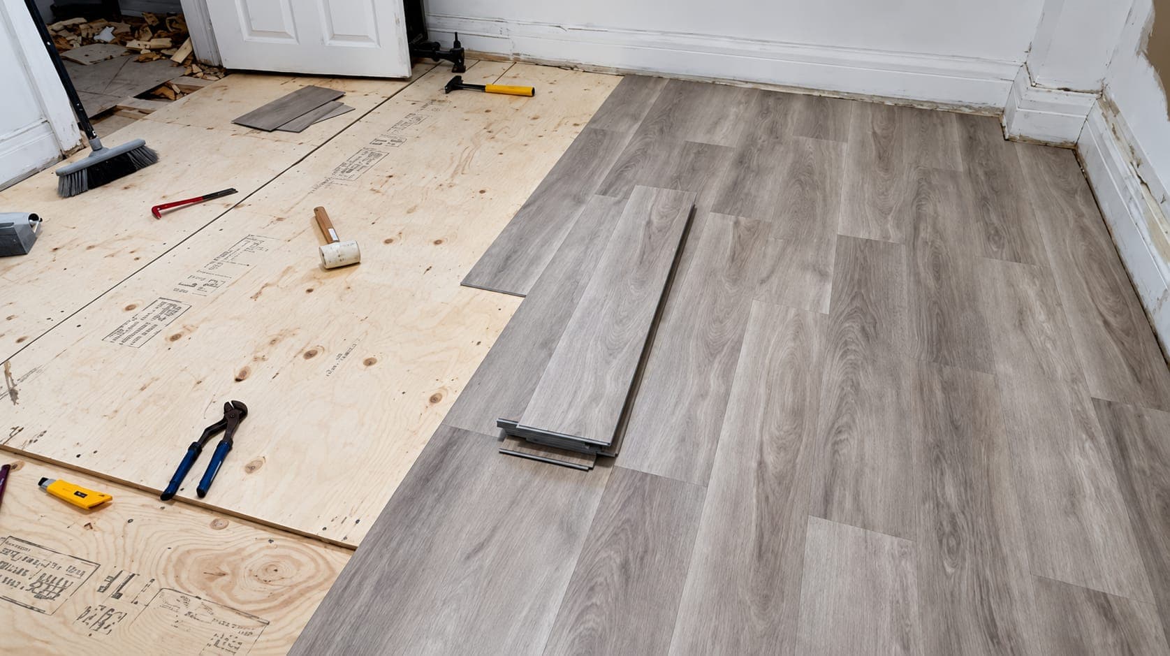 Gray wood-look vinyl plank flooring partly installed over a new plywood subfloor, with hand tools and loose planks