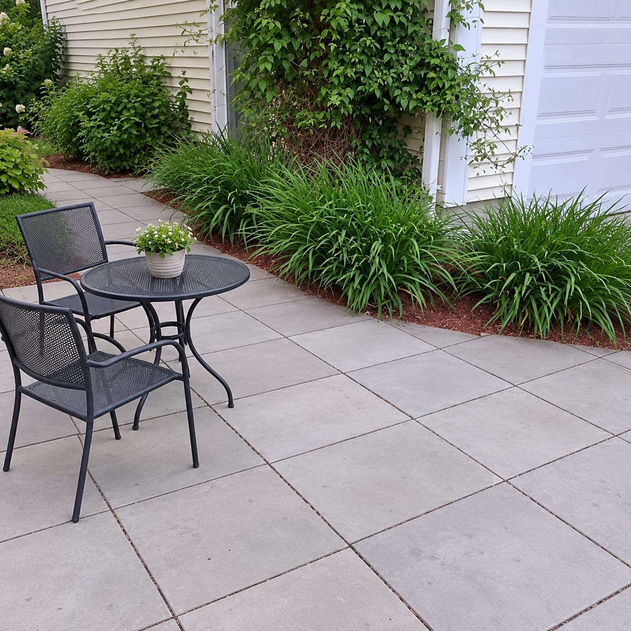 Patio of large square gray concrete pavers with a black metal bistro set next to a mulched planting bed