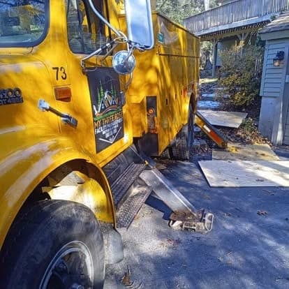 Yellow service truck with the company logo on its door, parked beside a gray house with plywood on the ground