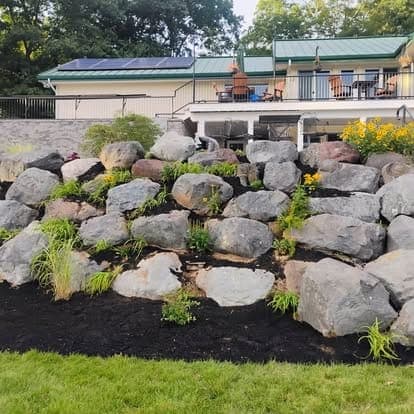 Stacked boulder retaining wall planted with yellow flowers and leafy perennials in dark mulch, below a green-roofed house