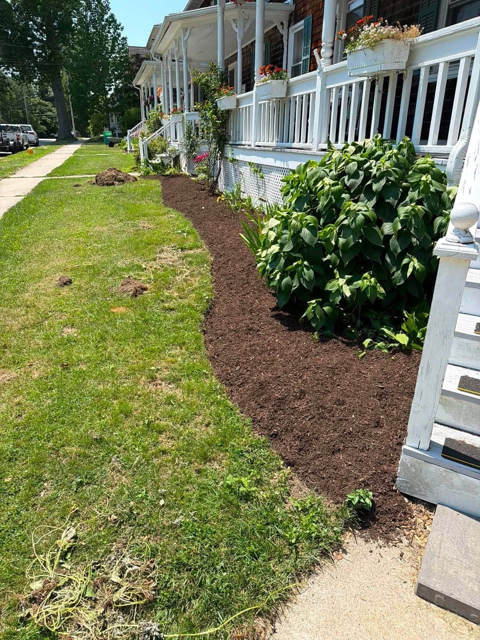Newly edged and mulched bed curving along a long white porch, with pulled sod clumps on the lawn by a sidewalk