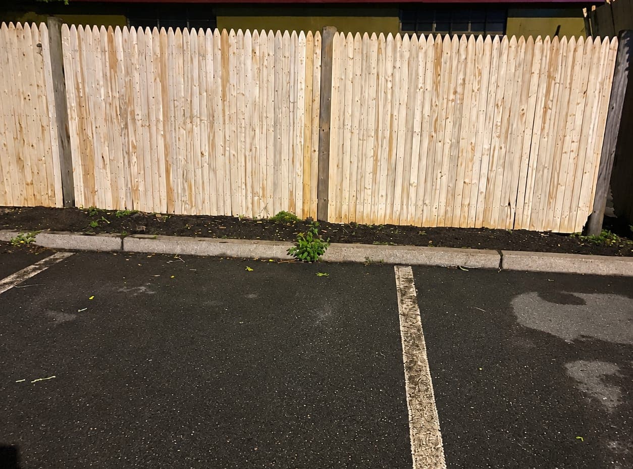 Row of new pale stockade pickets behind a fresh soil strip, concrete curb and white-striped parking spaces