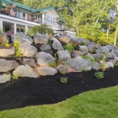 Boulder retaining wall on a black-mulched slope with new plantings, a metal stair railing and a building above