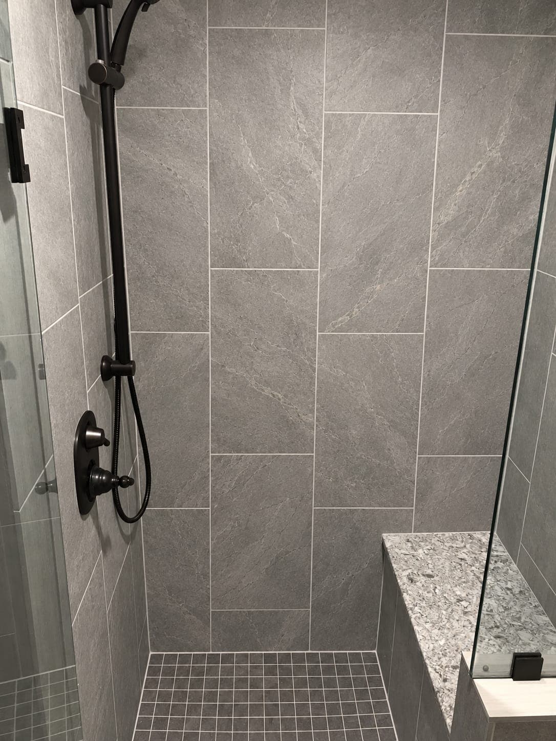 Walk-in shower with large gray stone-look wall tiles, a small square mosaic floor, a built-in bench and dark bronze fixtures
