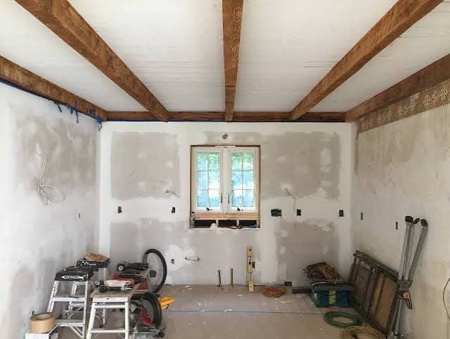 Room mid-renovation with patched drywall, exposed wood ceiling beams, a small window and power tools on the floor