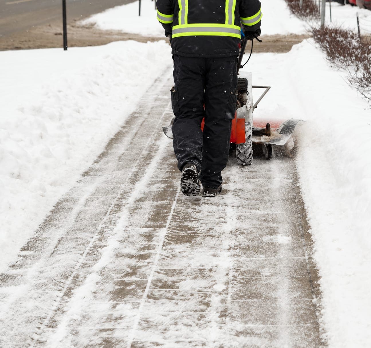 Worker in a high-visibility jacket clearing a snowy paver sidewalk with a walk-behind power broom between snowbanks