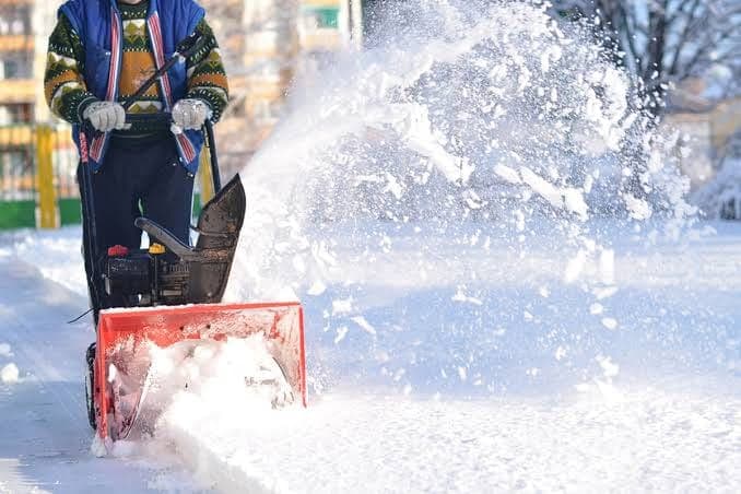 Person pushing a red walk-behind snow blower along a snowy walkway as it throws a plume of snow to the side