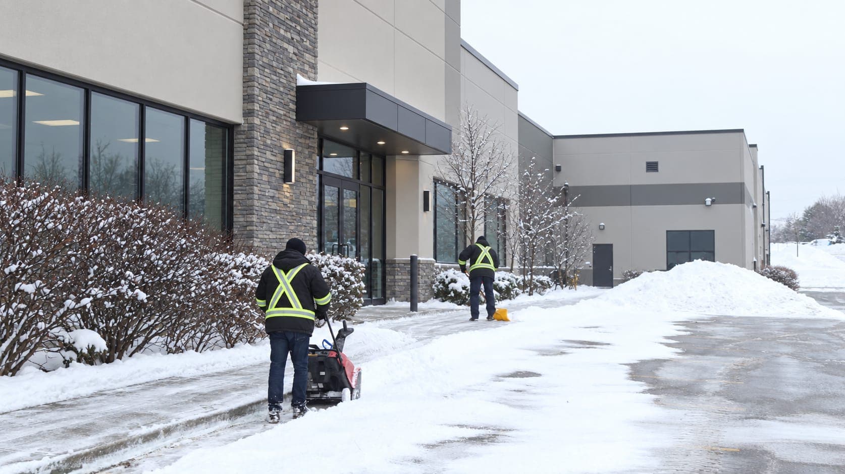 Two workers in reflective jackets clearing a snowy commercial sidewalk with a snow blower and a snow shovel